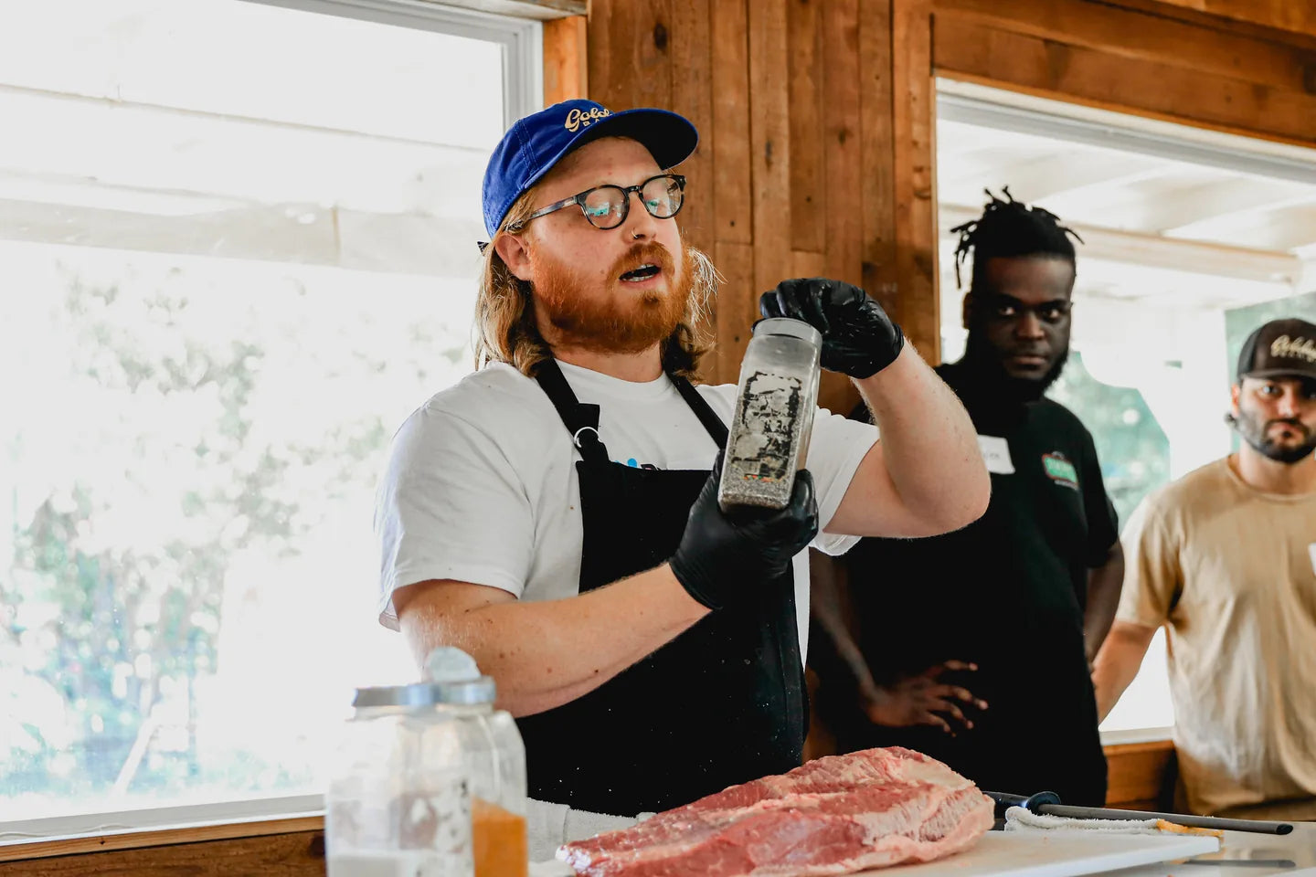 A Goldee's pitmaster holding up a jar of rub as he talks the class through it, a raw brisket on the table in front of him