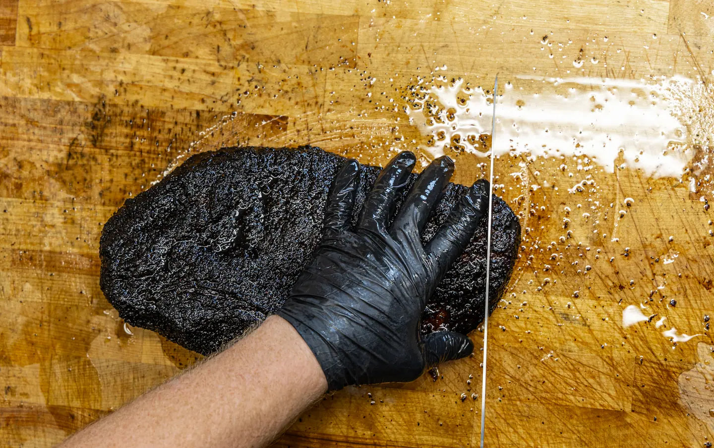 A gloved hand holding a whole brisket steady on the block as the slicing knife starts the cut