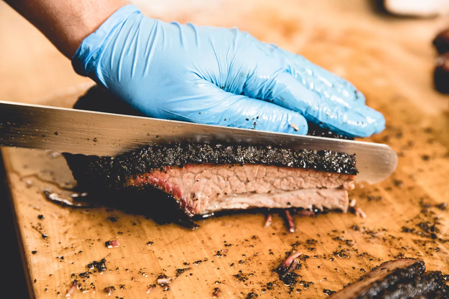 A slicing knife cutting through a brisket slice on the block, bark and smoke ring in view