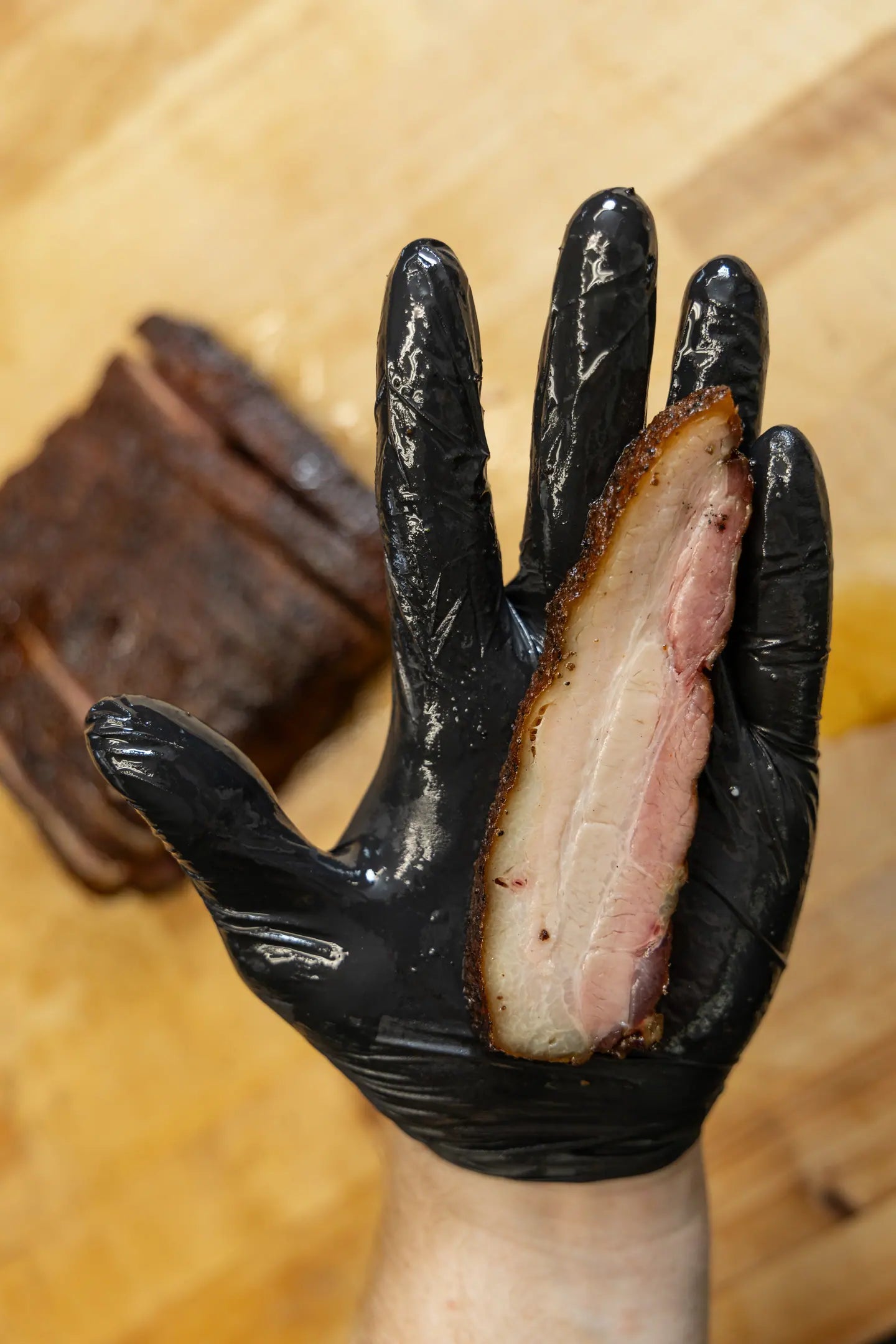 A thick slice of smoked pork belly held up in a gloved hand, its layers visible