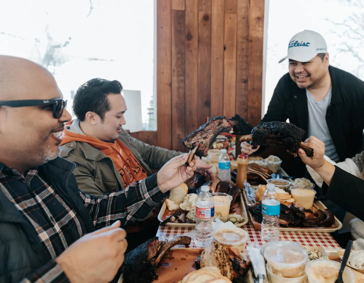A table mid-meal, trays of barbecue and sides between friends