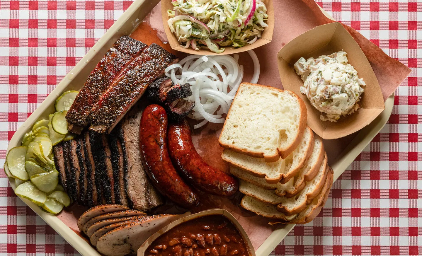 A tray of sliced brisket, sausage links, white bread, pickles, slaw and beans on red gingham paper