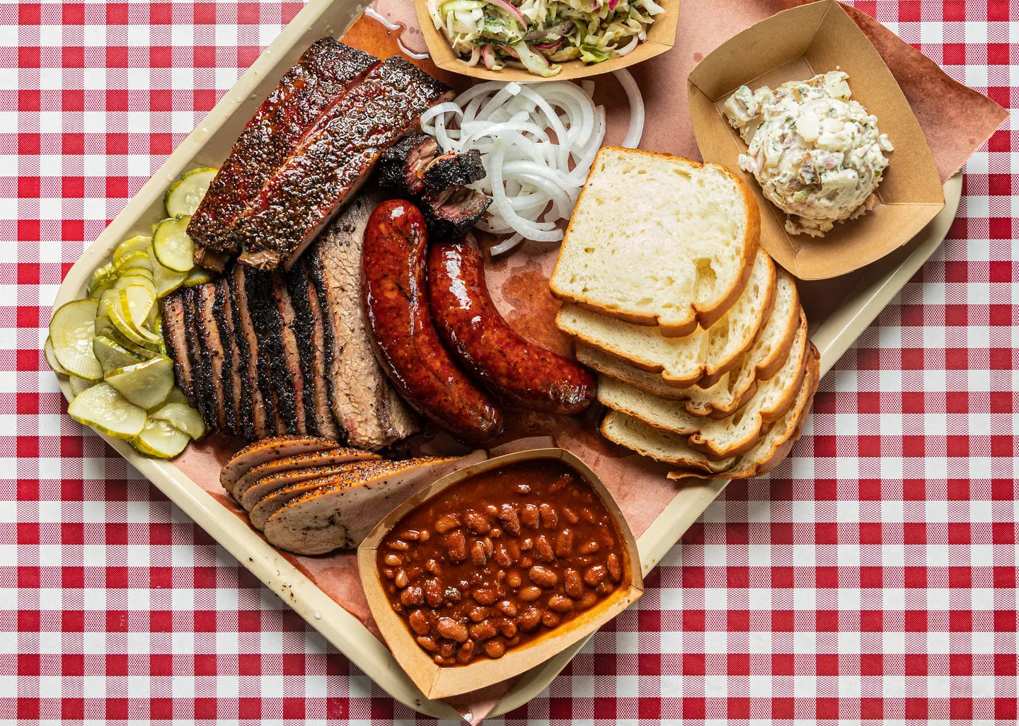 A tray of brisket, sausage, bread and sides on red gingham, photographed from above