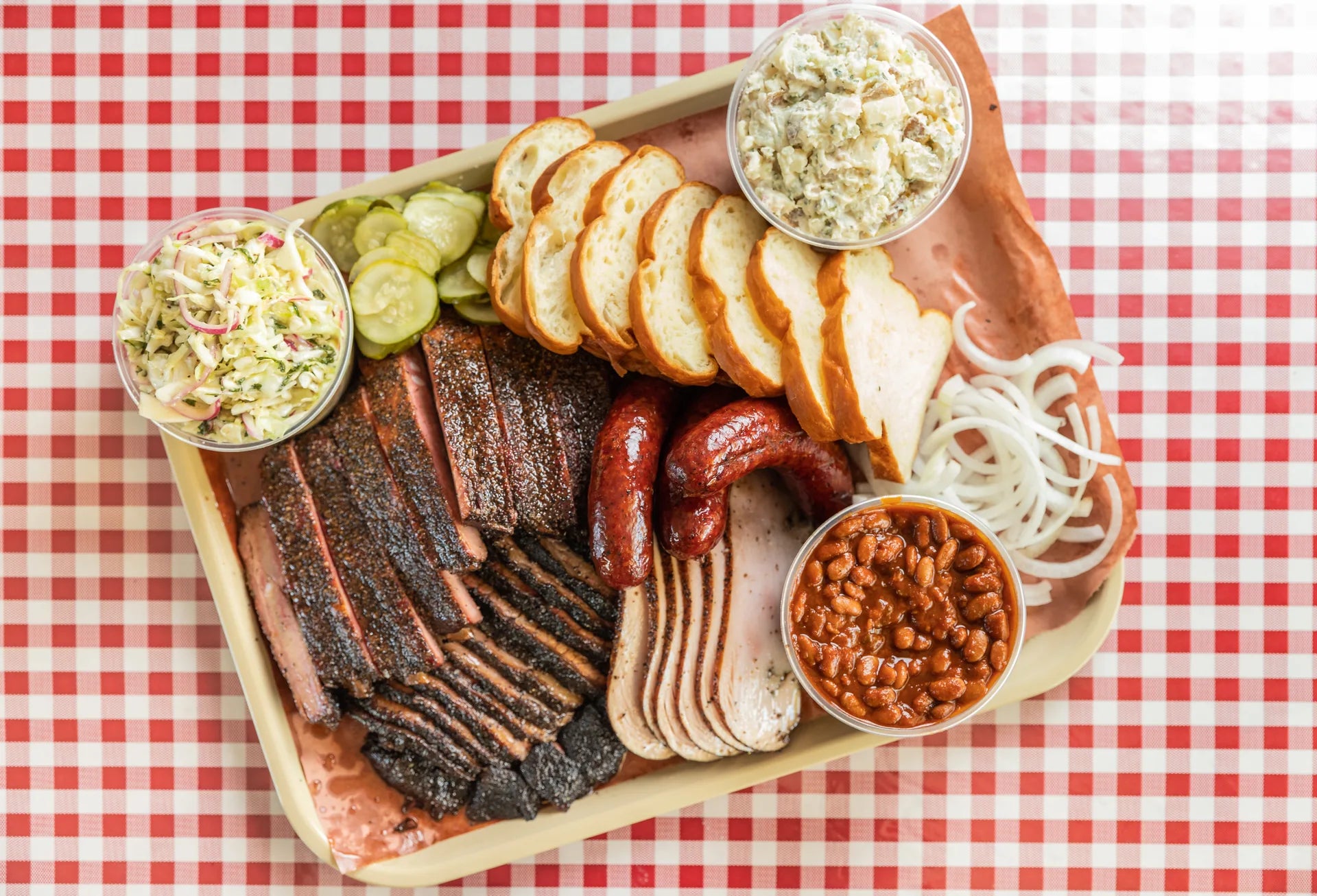 A full tray of brisket, turkey, sausage, bread and sides photographed straight down on red gingham paper